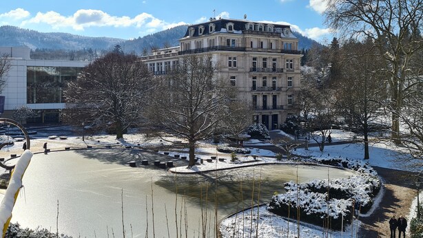 Un grand bâtiment historique, doté de plusieurs étages et d’un balcon imposant, entouré d’arbres couverts de neige et d’un étang gelé.