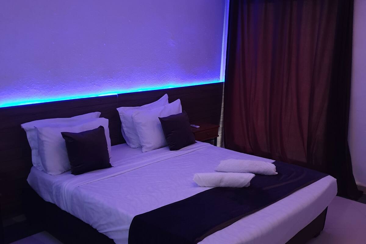 A neatly made bed with white and dark pillows, a dark headboard, and a blue ambient light strip.
