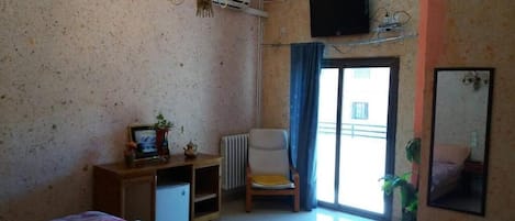 Quarto com cama, uma cadeira, uma mesinha com porta-retrato, radiador, televisão fixada na parede e janela com cortinas.