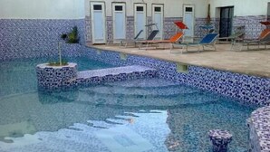 Una piscina con paredes de azulejos y escalones que bajan al agua. Se ven sillones de descanso y una puerta al fondo.
