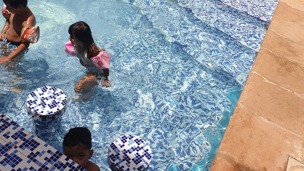 A swimming pool with blue and white mosaic tiles, a palm tree, and a few people swimming and playing. - Le Privilege Hotel (Oran)