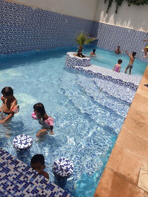 Uma piscina com azulejos brancos e azuis, uma palmeira e algumas pessoas nadando e brincando.