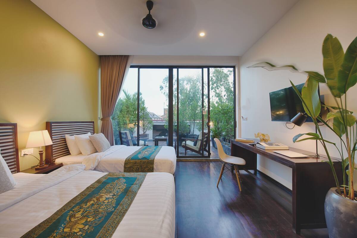 A hotel room with two beds, a desk, a chair, a TV, and a large window with a view of trees and buildings.