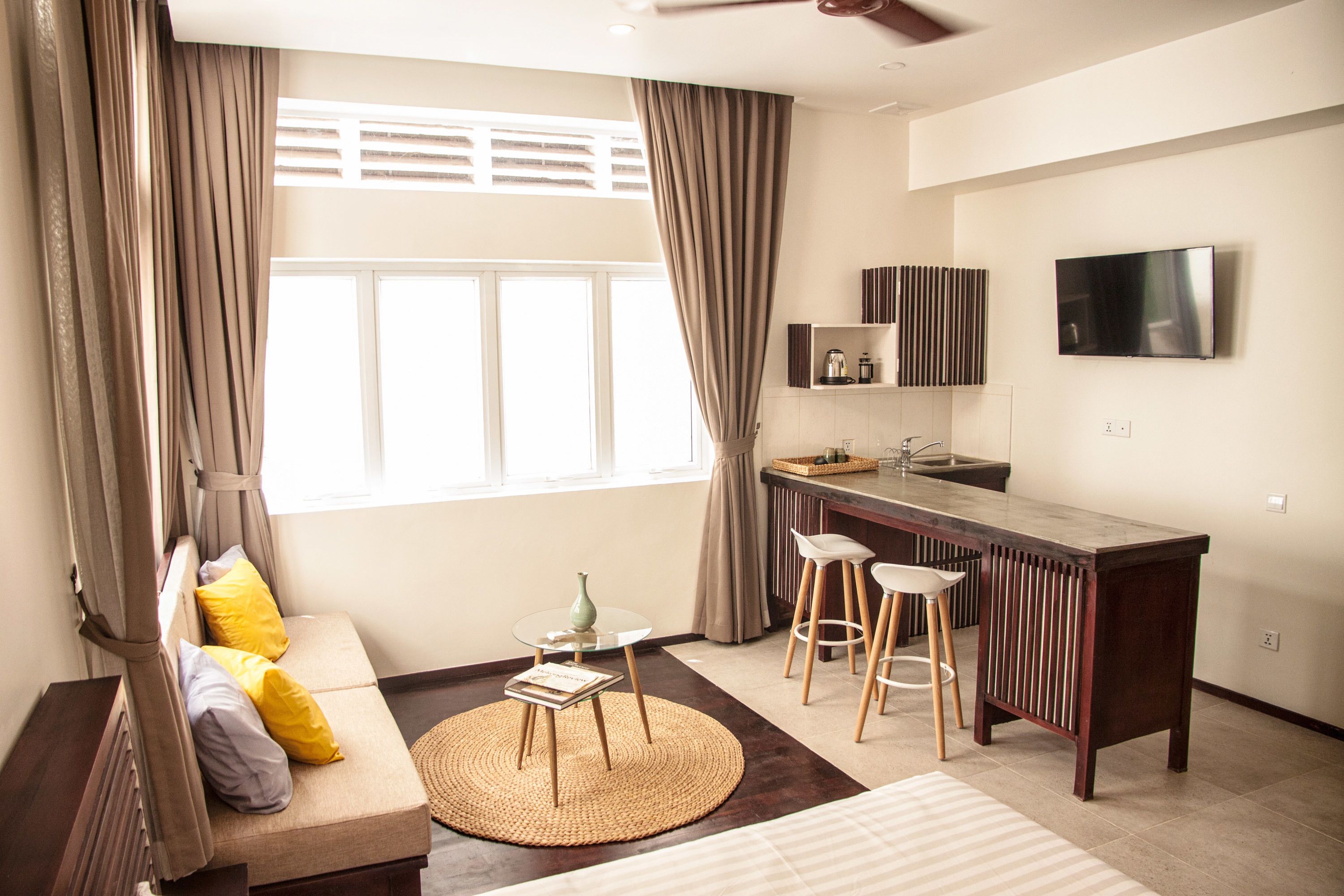 A modern hotel room with a sofa, a small round table, a bar area with stools, a TV mounted on the wall, and a large window with curtains.