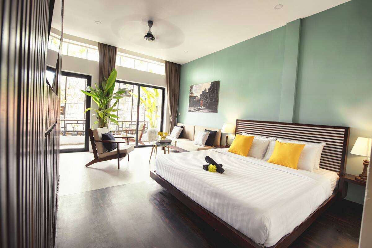 A hotel room with a large bed, a sitting area with a chair and table, a potted plant, and a view through the window.