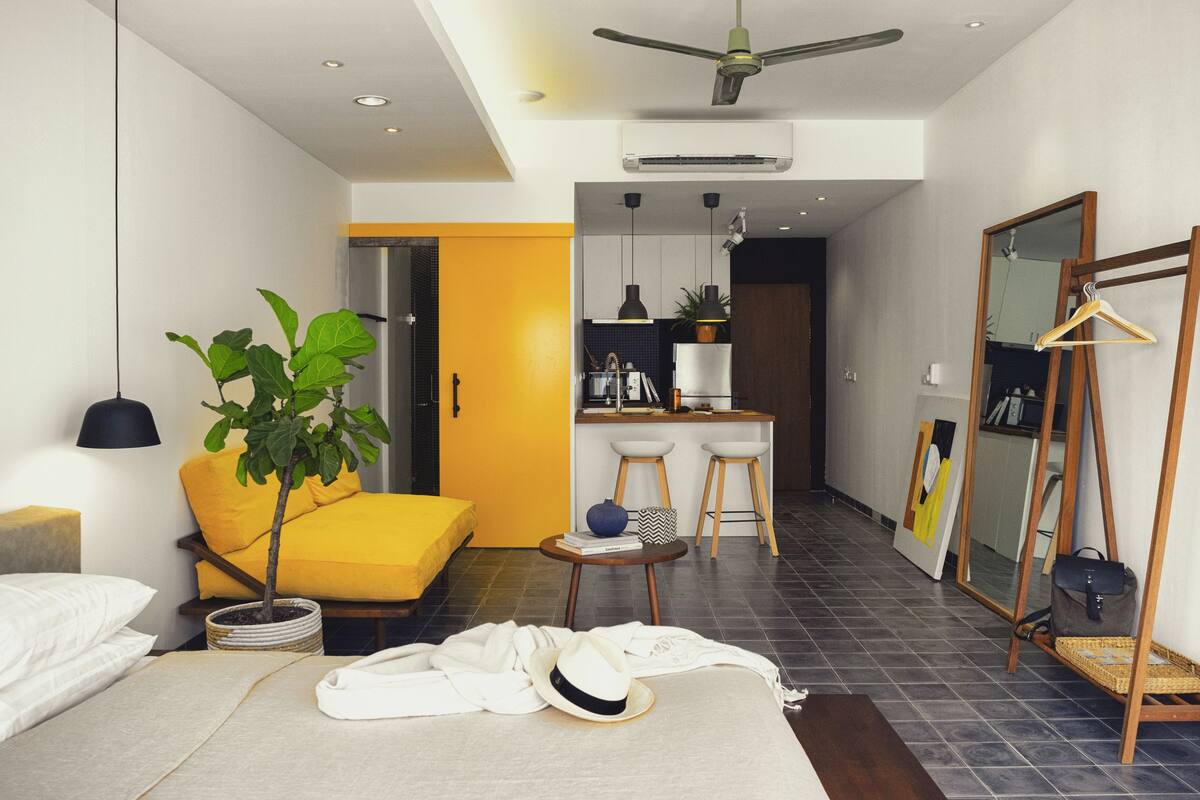 A modern hotel room with a bed, a small table with a plant, a yellow sofa, and a kitchenette in the background.