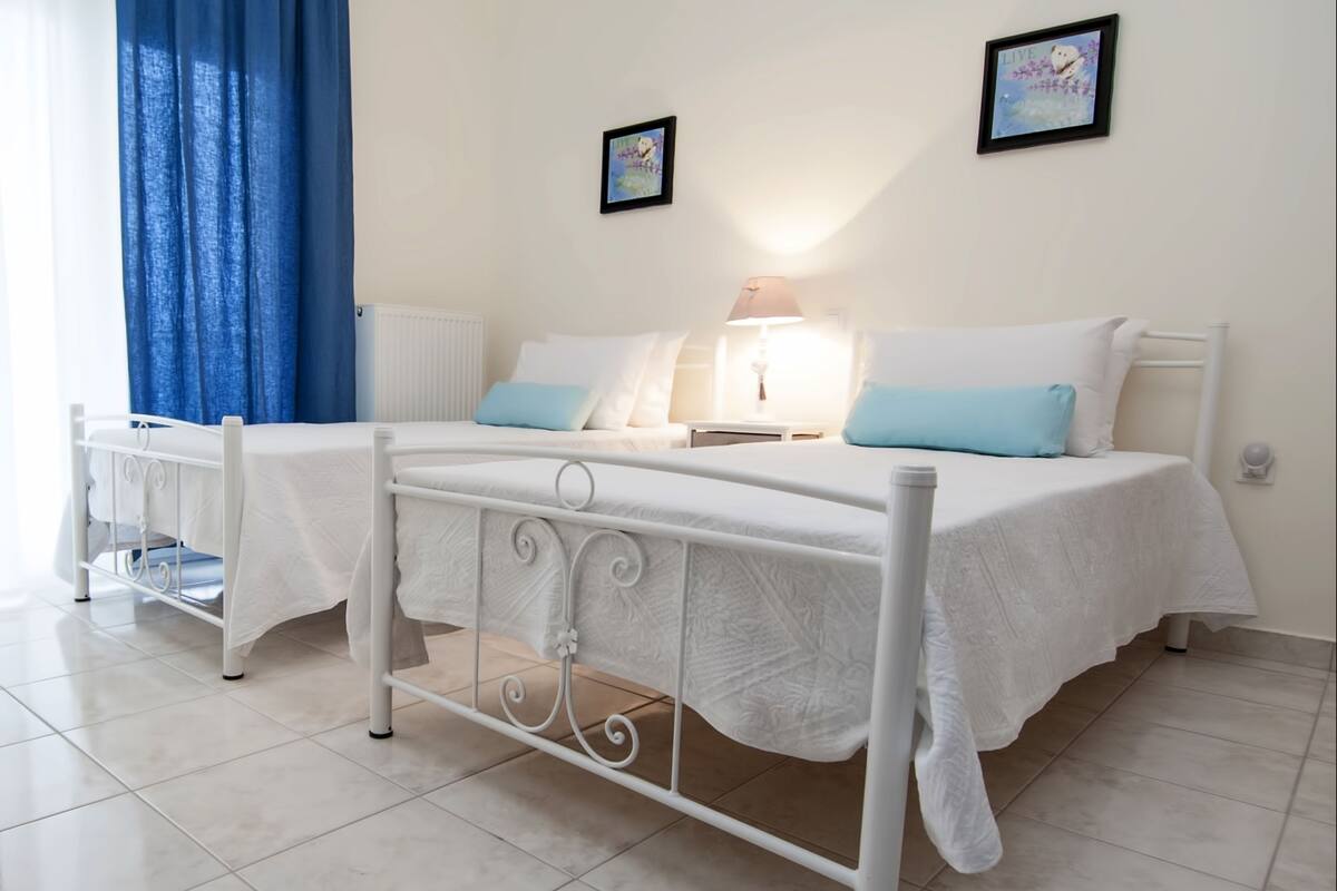 A double bed with white bedding and blue pillows, a bedside table with a lamp, and a window with blue curtains.