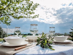 A table set for a meal with white plates, bowls, and wine glasses, surrounded by greenery and a clear sky. - Villa Elaia Mare (Komotini)