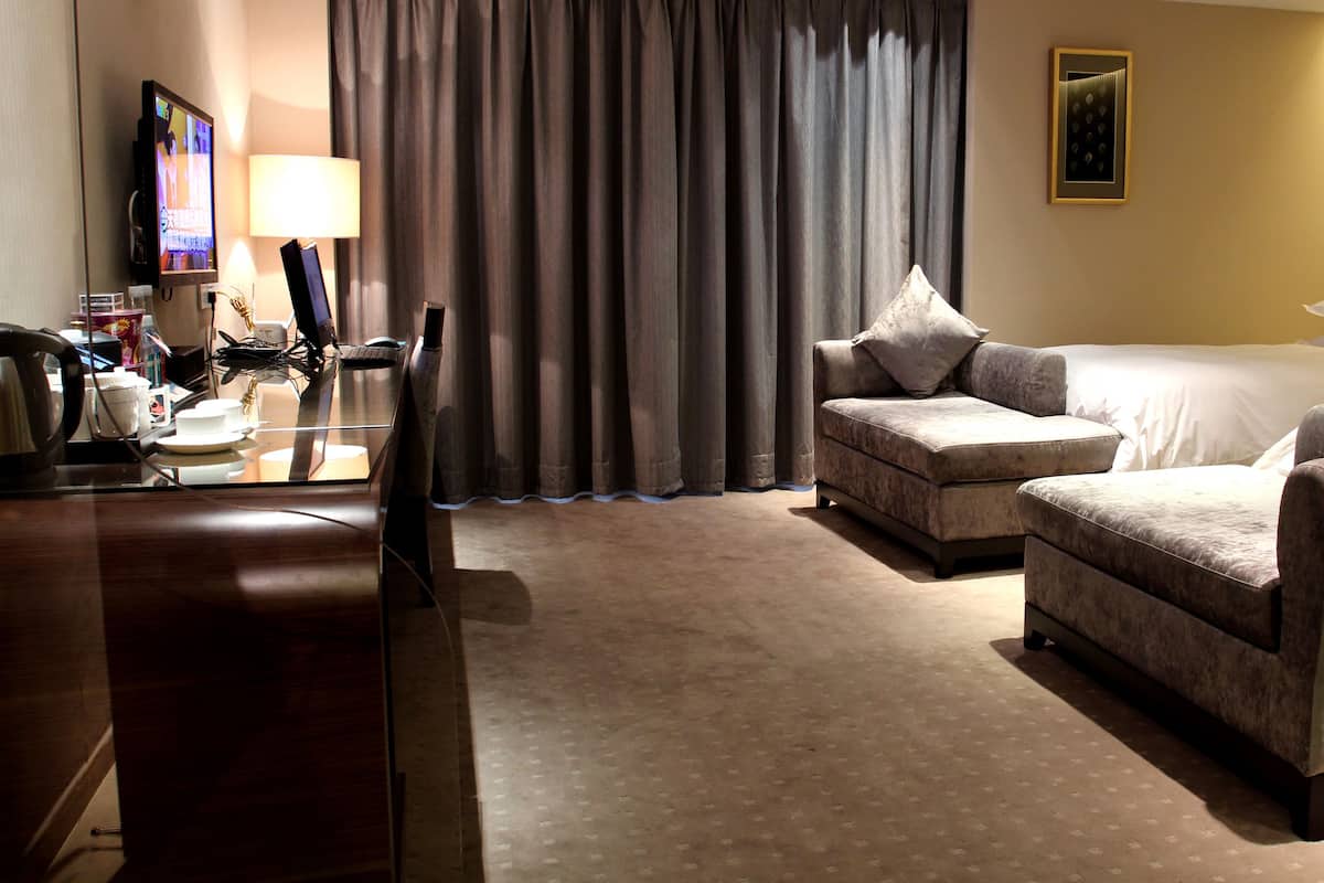 A hotel room with a bed, two armchairs, a desk with a computer, a TV, and a large window with curtains.