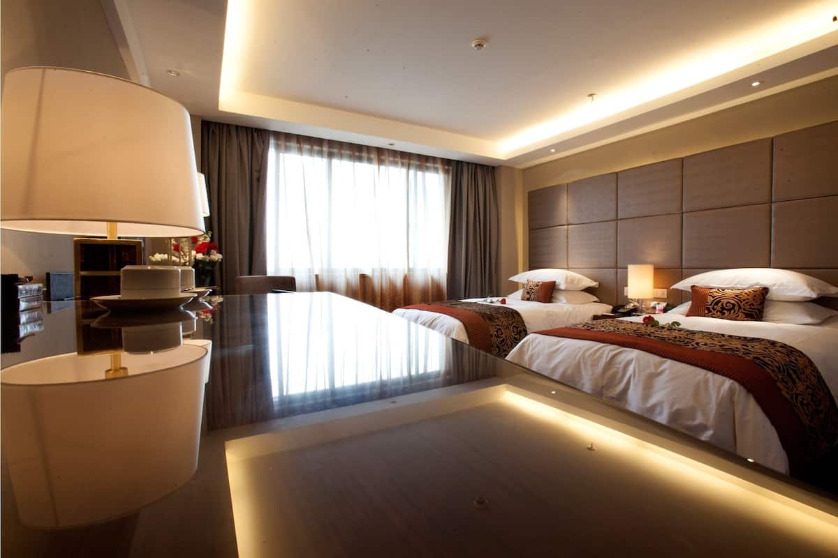 A modern hotel room with two beds, a large desk, a lamp, and a view of the outdoors.