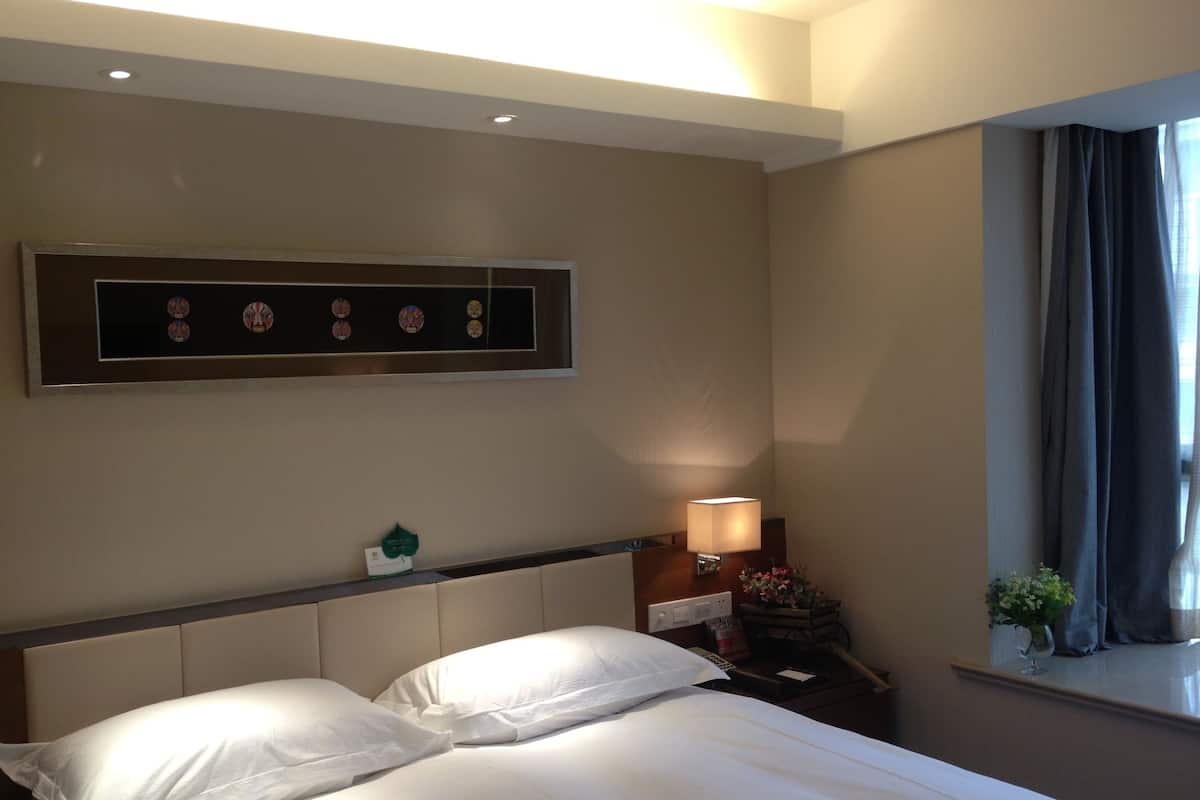 A hotel room with a bed, a nightstand with a lamp, a window with curtains, and a wall-mounted artwork.
