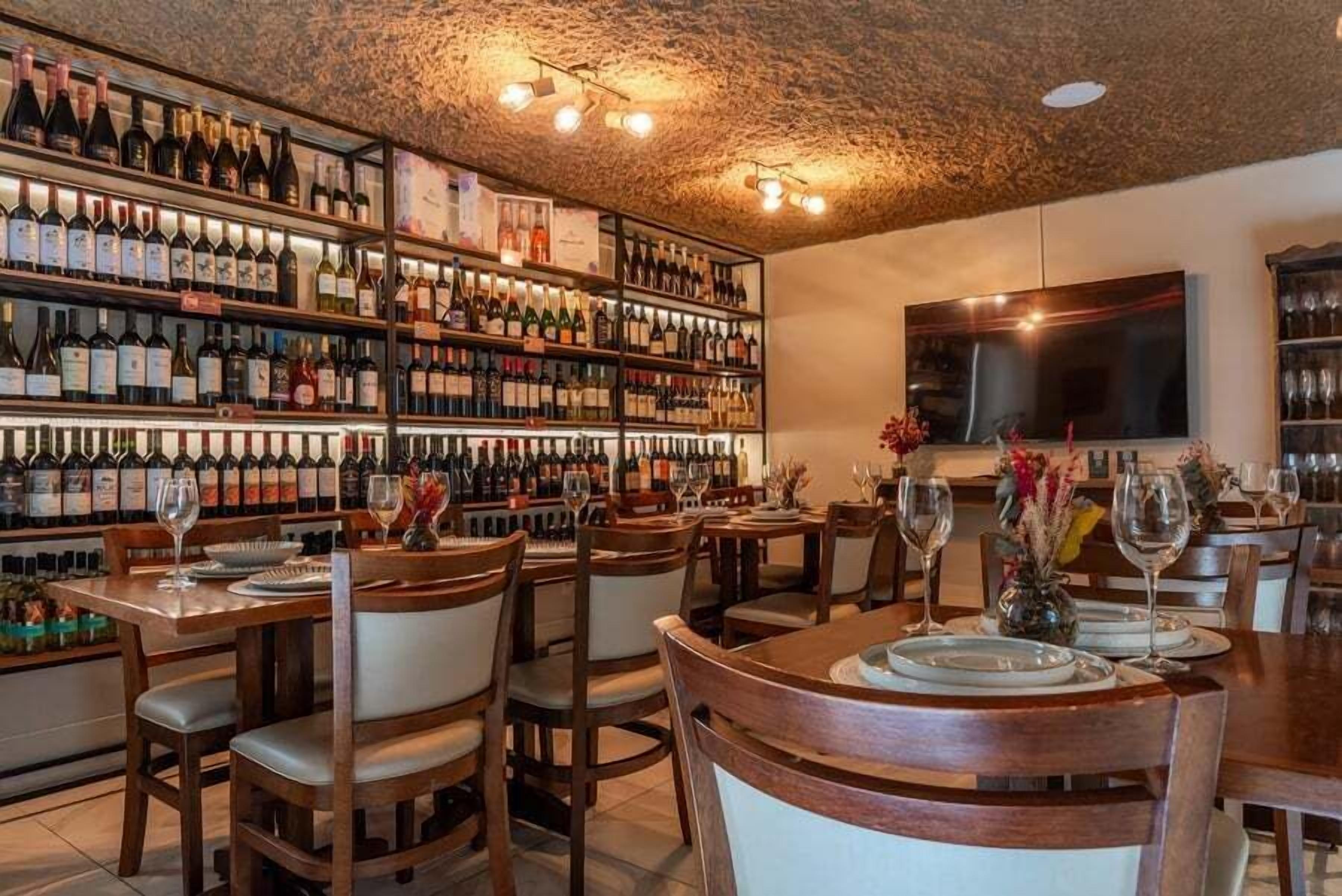 A restaurant with multiple tables, each set with wine glasses and plates, and a wall of wine bottles behind the tables.