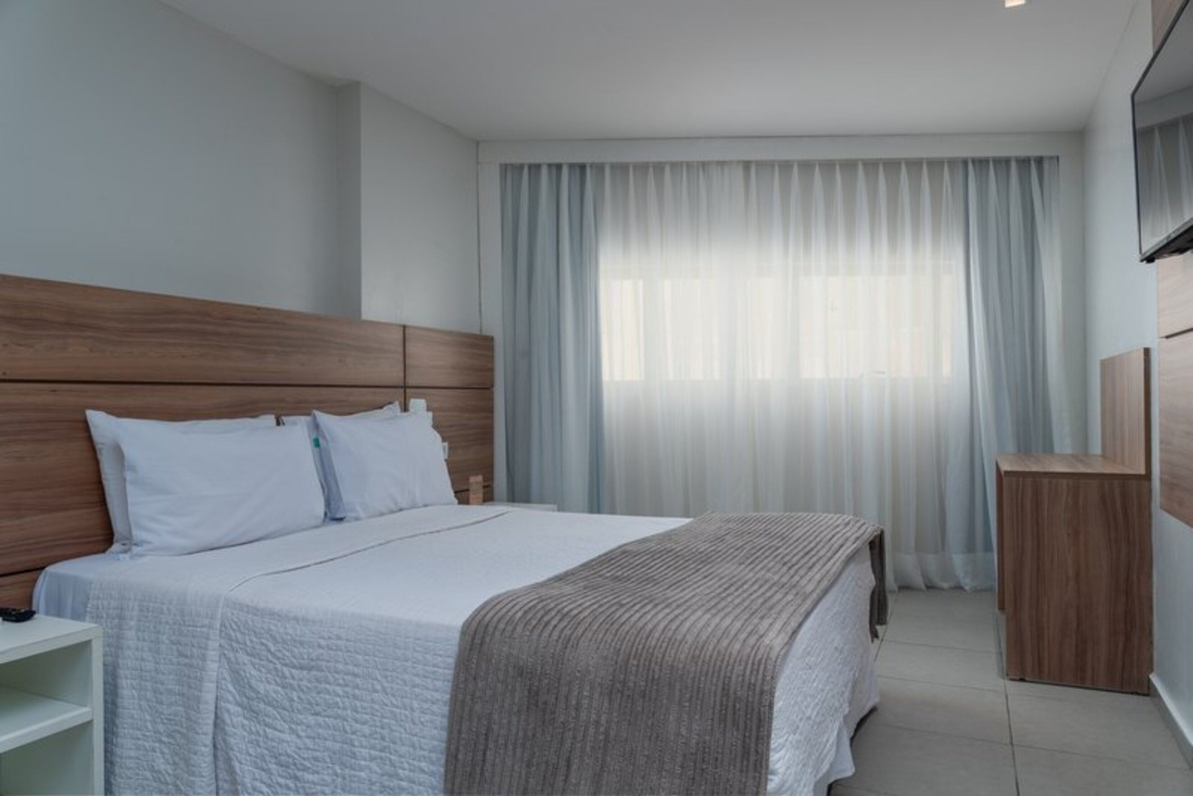 Una habitación de hotel moderna con una cama grande, cabecera de madera, ropa de cama blanca y un ventanal con cortinas de gasa.
