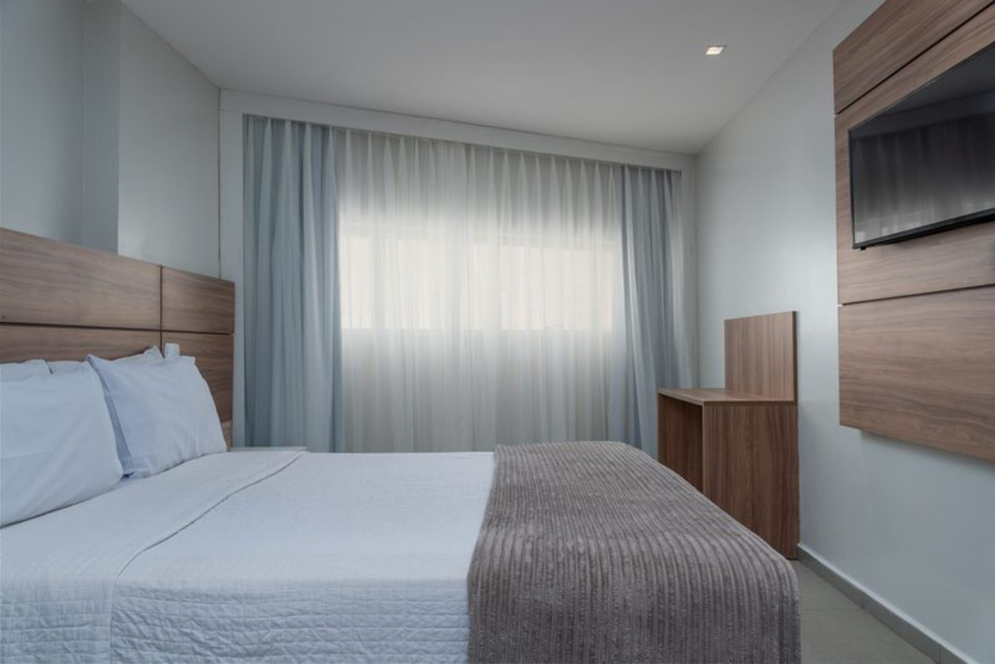 Una habitación de hotel moderna con una cama grande, un televisor montado en la pared y una ventana con cortinas traslúcidas.