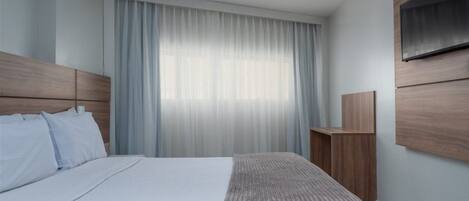 Una habitación de hotel moderna con una cama grande, un televisor montado en la pared y una ventana con cortinas traslúcidas.