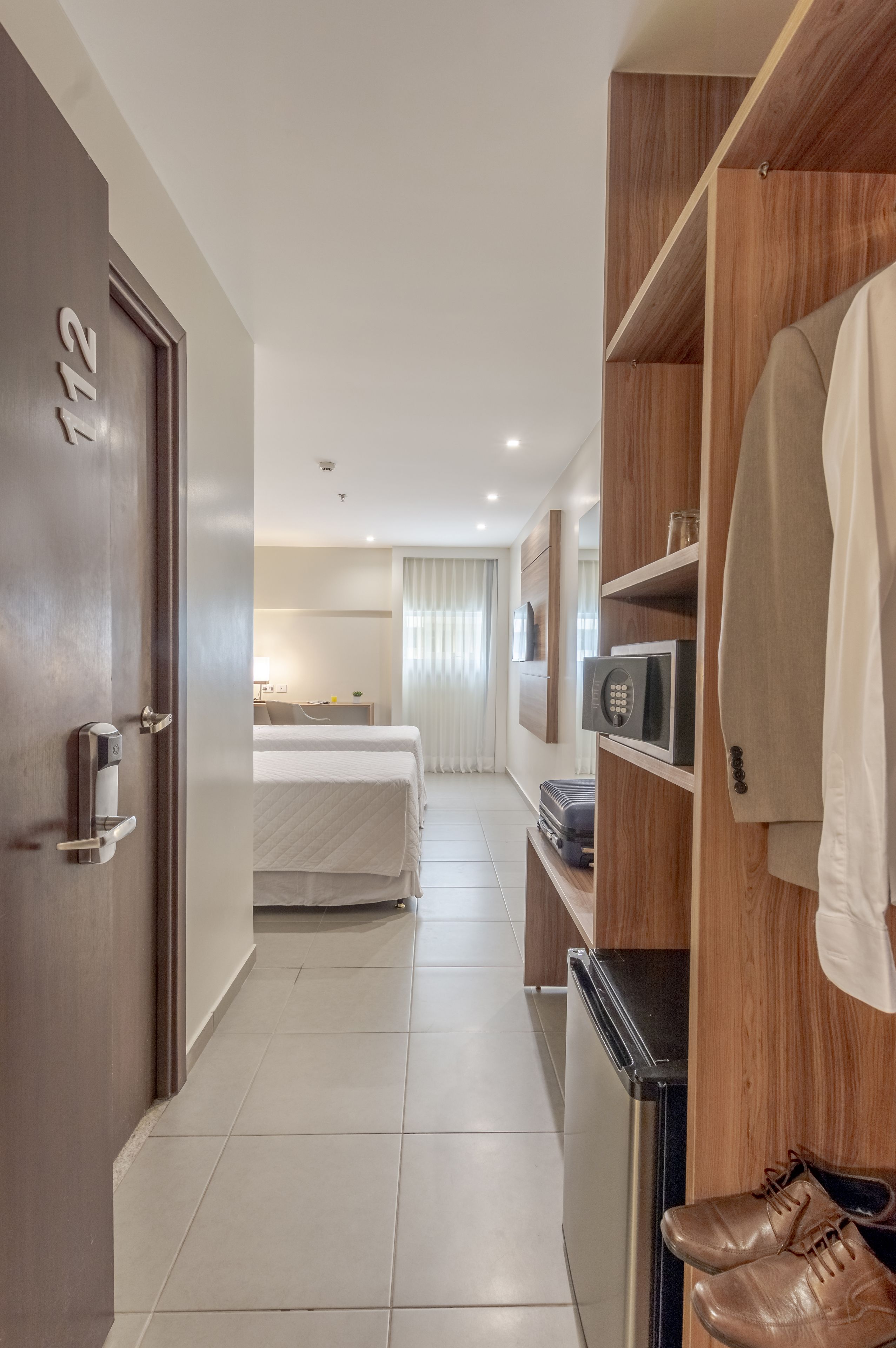 A hotel hallway with a door labeled "112", a bed, a microwave, and a shoe rack.