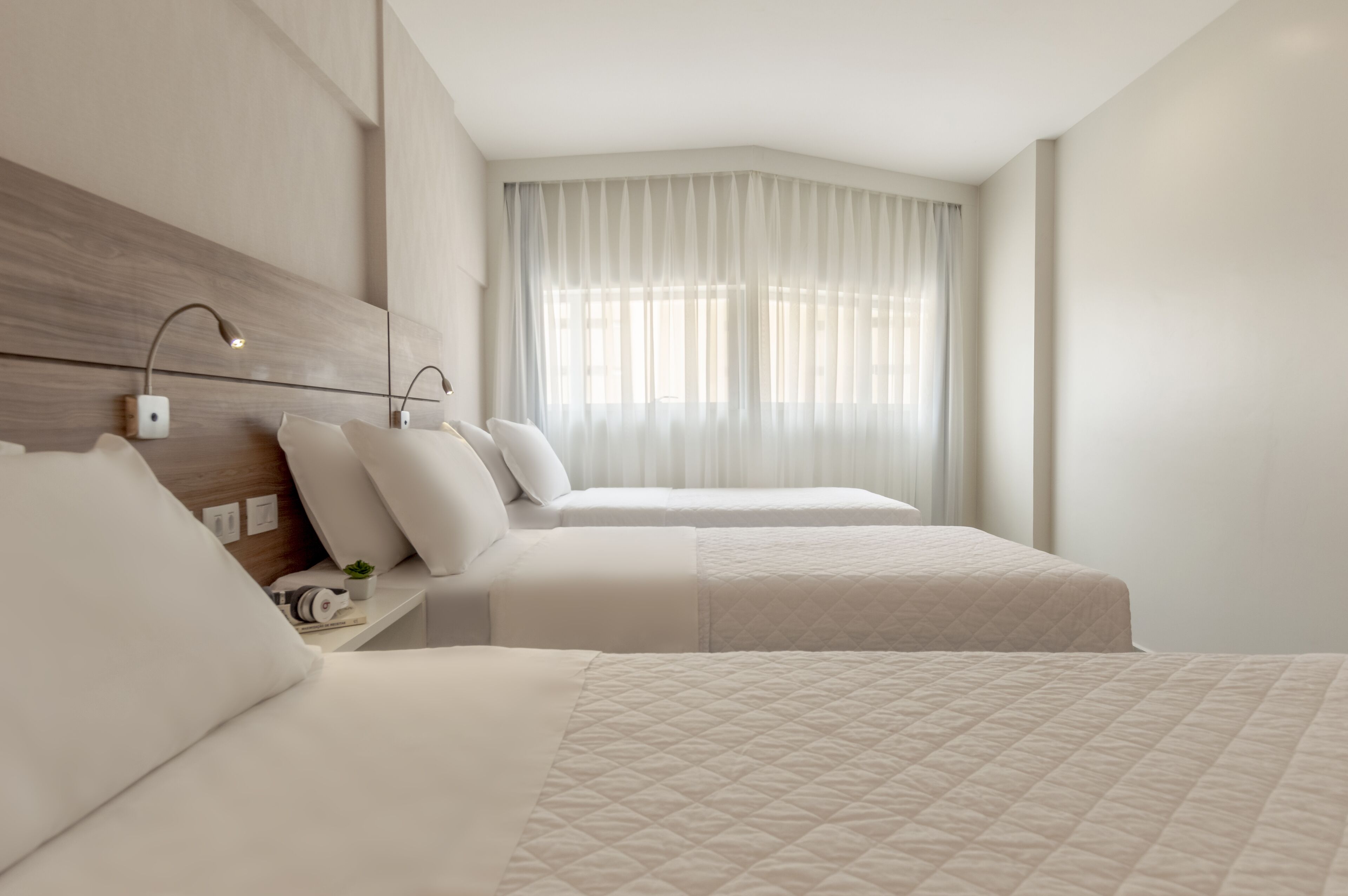 A hotel room with two beds, a wooden headboard, a bedside table with a lamp, and a large window with sheer curtains.