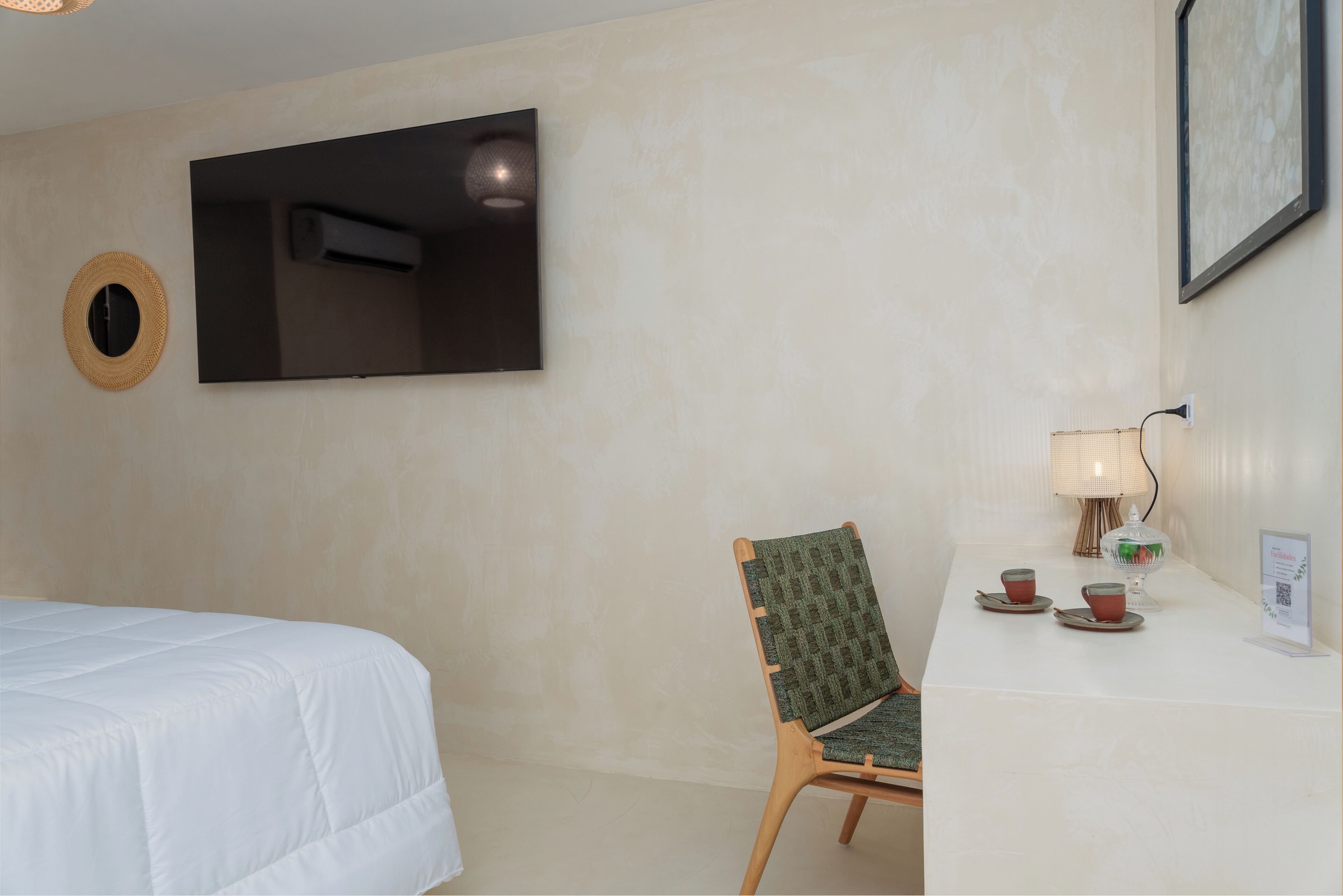 A hotel room with a bed, a chair, a table with cups and a lamp, and a TV mounted on the wall.