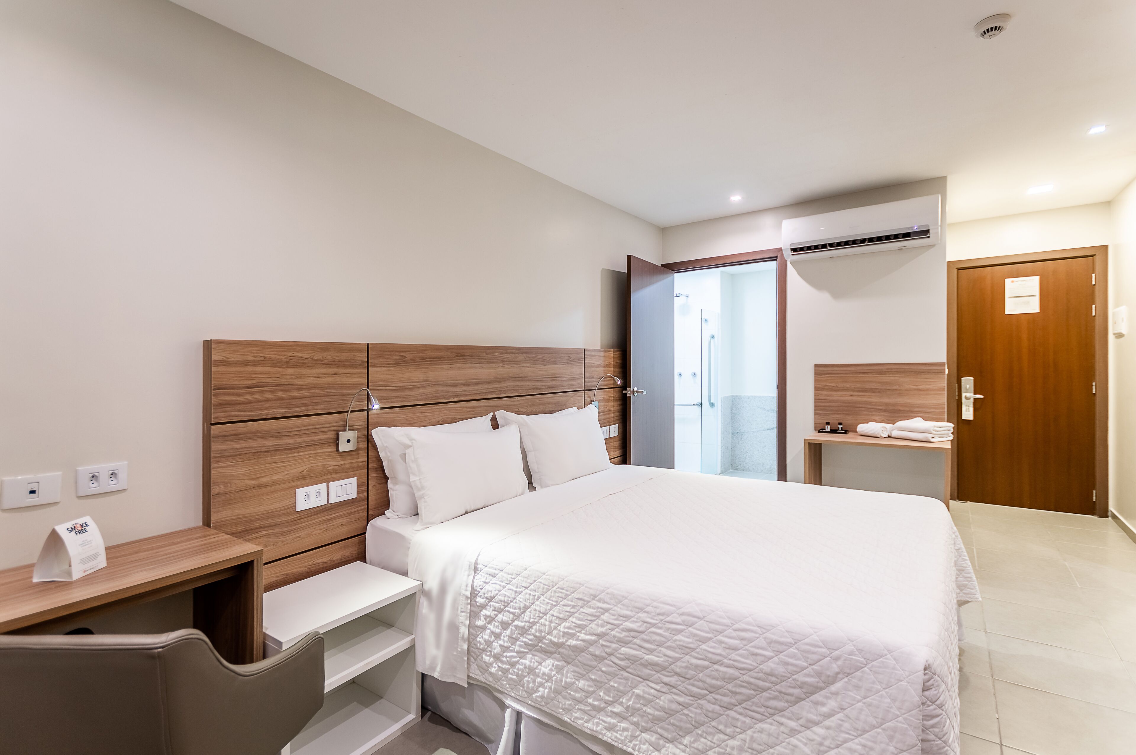 A modern hotel room with a wooden headboard, a large bed with a quilted white duvet, a bedside table with a lamp, and a small desk with a chair.