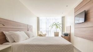 A modern hotel room with a large bed, a seating area, a TV mounted on the wall, and a potted plant.