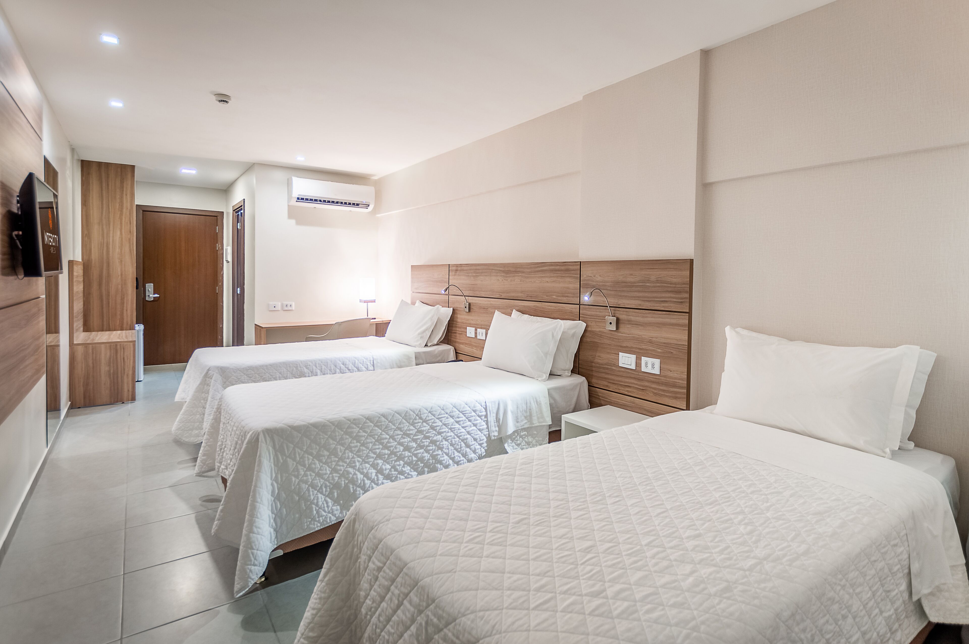 Habitación de hotel con tres camas, cabecera de madera y televisor de pantalla plana en la pared.