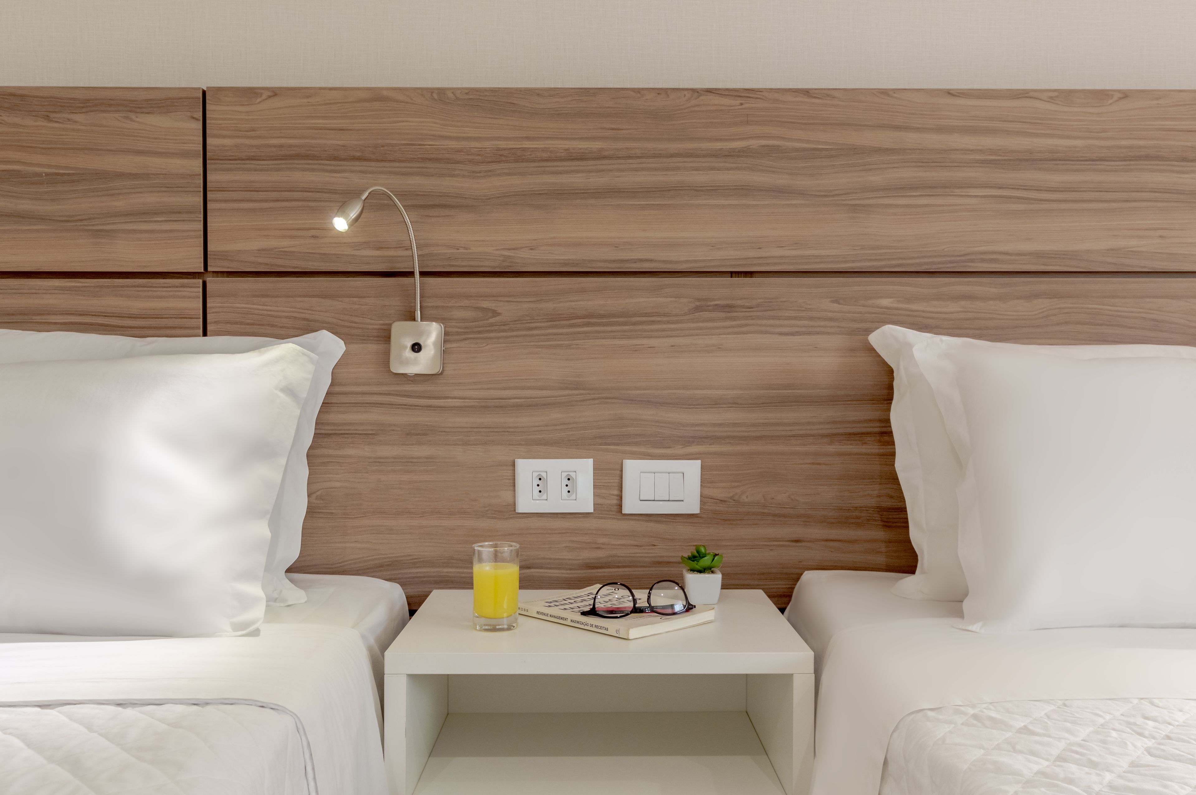 A hotel room with two beds, a bedside table with a lamp, a glass of orange juice, a pair of glasses, and a small potted plant.