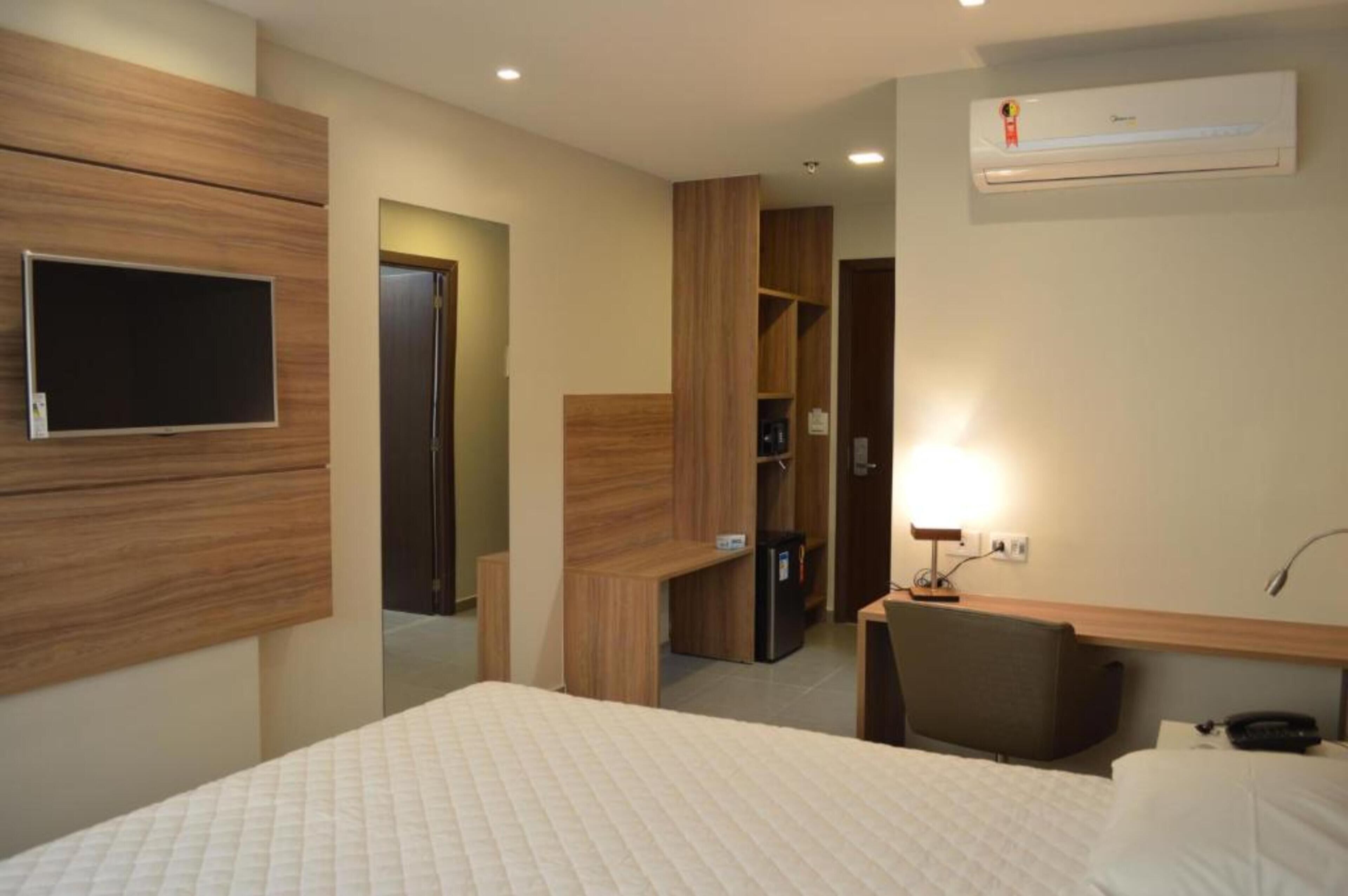 A hotel room with a bed, a desk, a chair, a TV, and an air conditioning unit.