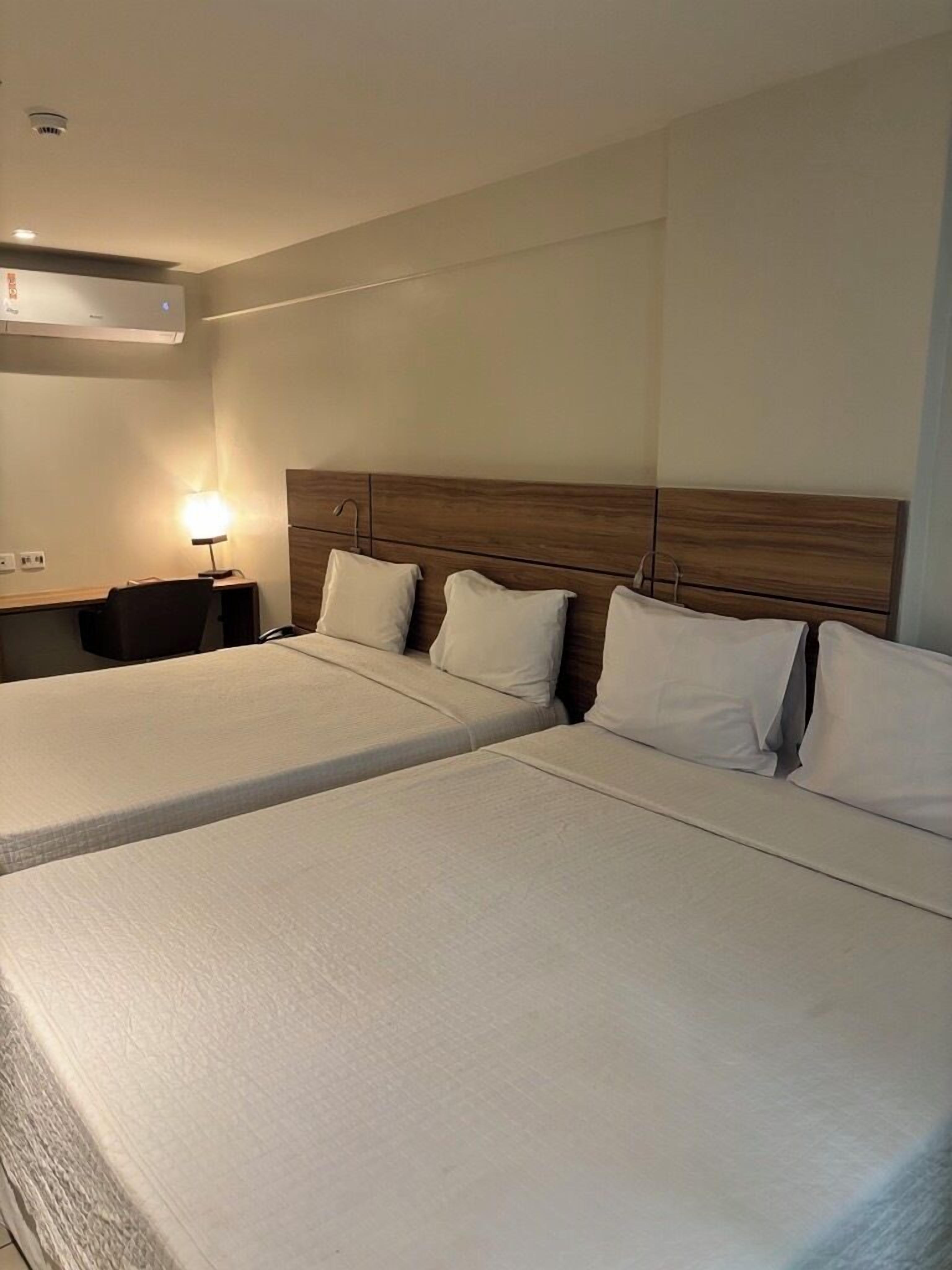 A hotel room with two beds, a wooden headboard, a desk with a lamp, and a wall-mounted air conditioning unit.