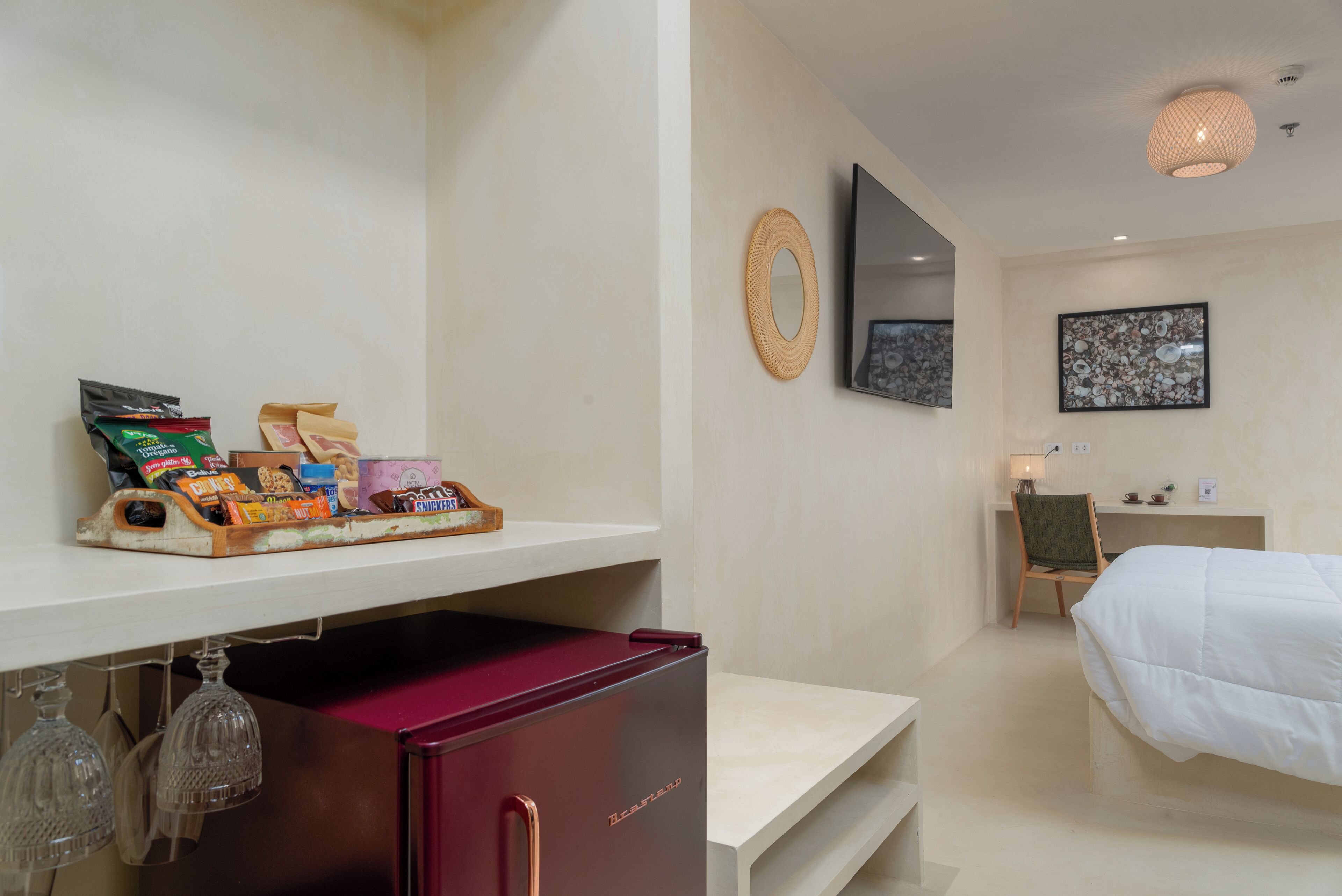 A hotel room with a bed, a desk, a shelf with snacks, a mirror, and a wall art piece.