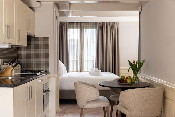 A compact hotel room with a kitchenette, a bed with rolled towels, a dining area with chairs, and a window with curtains.