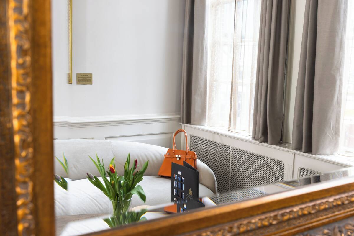 A hotel room with a bed, a window with curtains, a radiator, a plant, and a handbag on the bed.