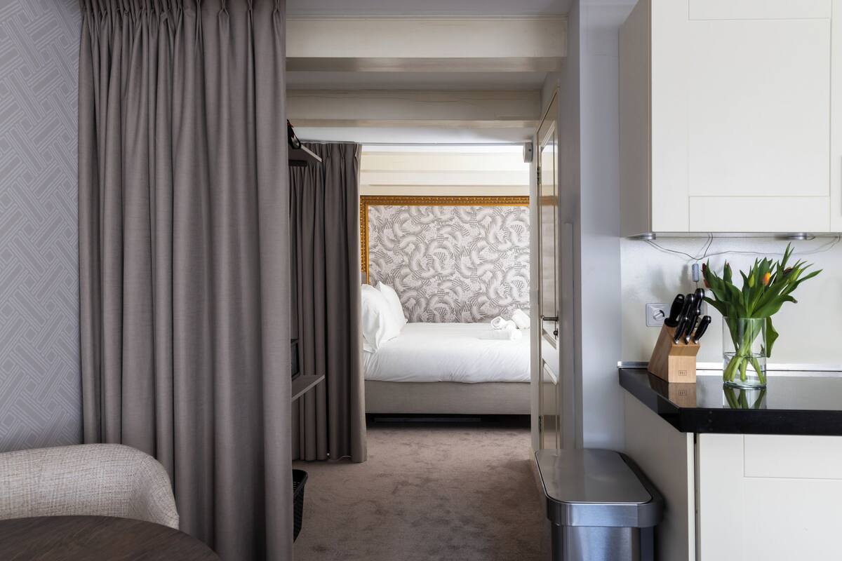 A hotel room with a bed, a bedside table with a vase and a knife block, and a view into another room with a patterned wall.