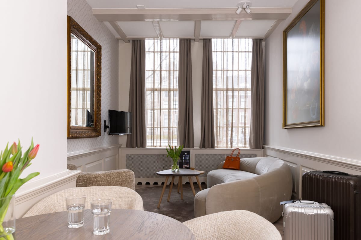 A hotel room with a seating area, a table with glasses, a vase with flowers, a television, and a large window with curtains.