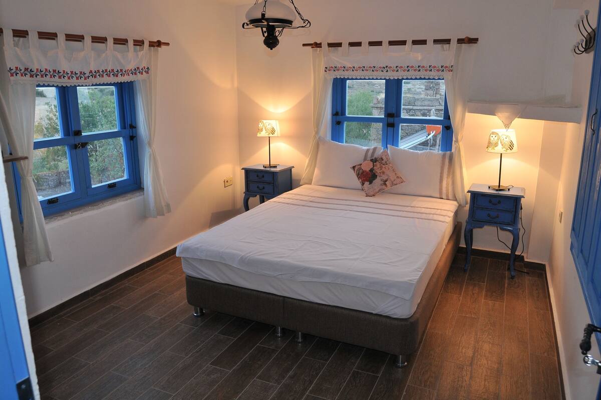 A bedroom with a large bed, two bedside tables with lamps, a window with blue shutters, and a wooden floor.