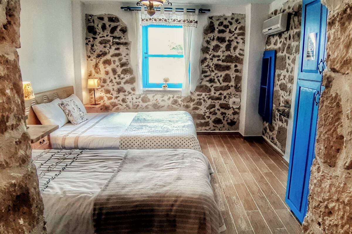 A room with two beds, a stone wall, a window with blue shutters, and a blue door.