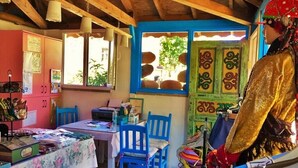 Quarto com teto de madeira, mesa com cadeiras, janela com vista para vegetação e porta colorida com desenhos decorativos.