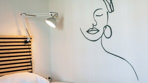 Une chambre à coucher avec un lit, une lampe fixée au mur, une serviette et une œuvre d’art murale représentant le profil d’une femme.
