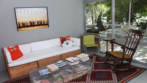 Un salon moderne avec un canapé blanc, une table basse en bois sur laquelle sont posés des livres, un tapis à motifs et une vue sur l’extérieur grâce à de grandes fenêtres.
