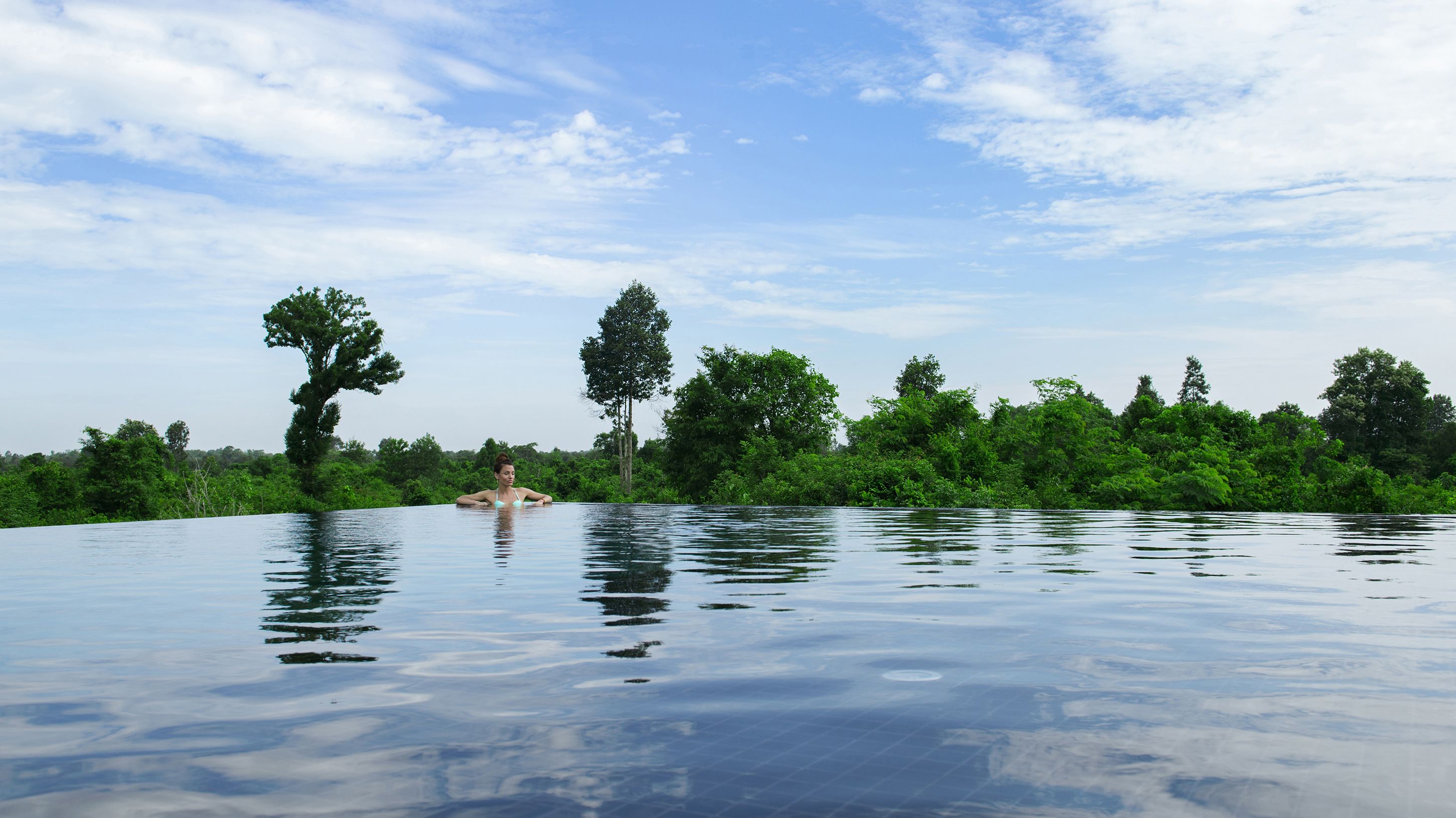 Una persona en una piscina infinita, con un cielo despejado y abundante vegetación al fondo.