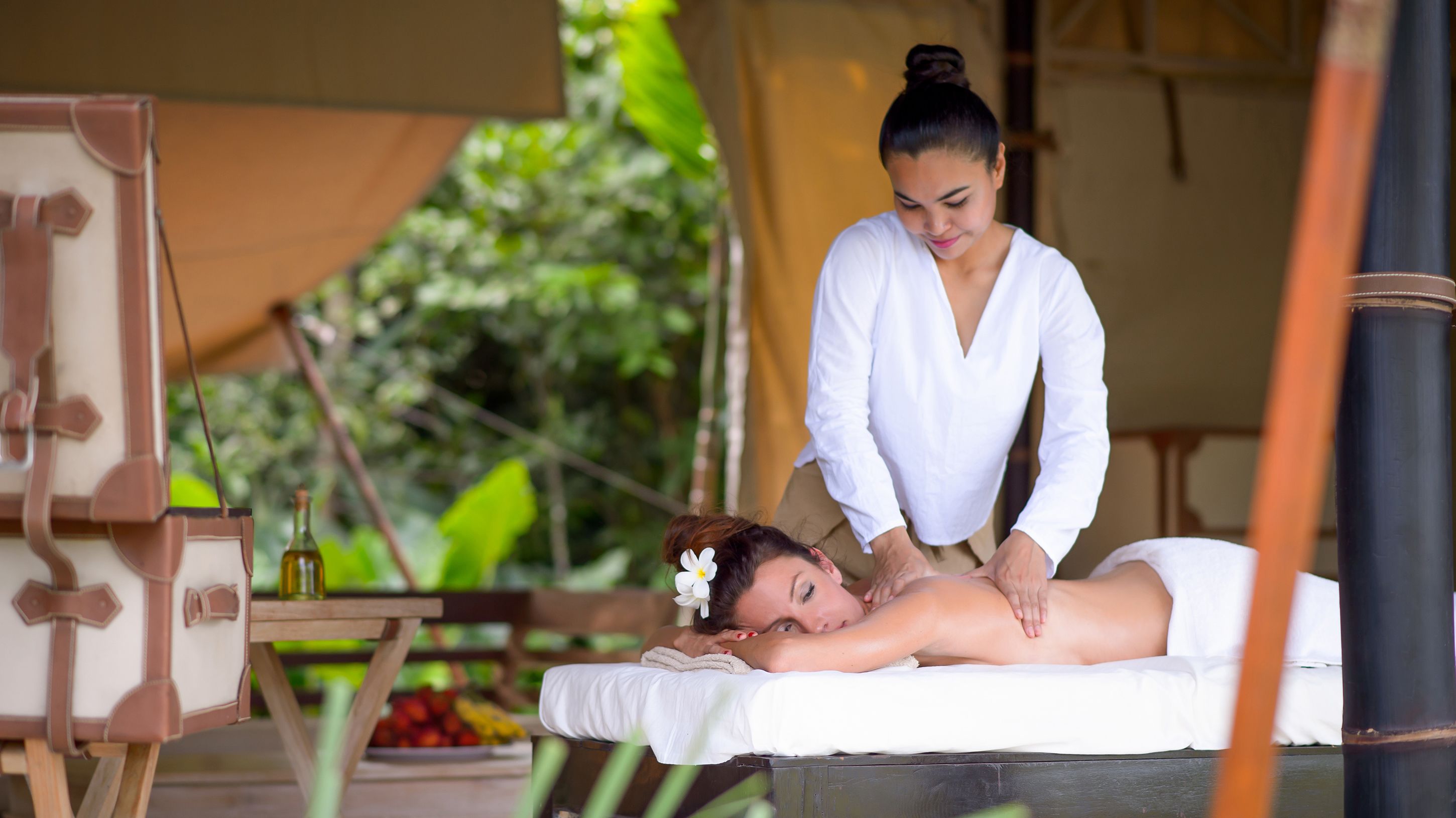 A massage therapy session in a tented area with a wooden table, a bottle of oil, and a bowl of fruit in the background.