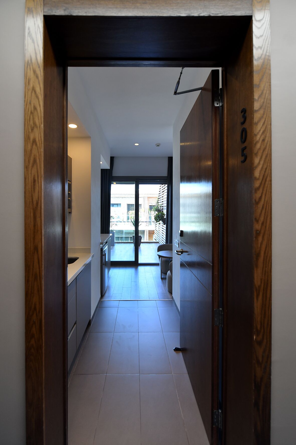 A modern hallway with a wooden door marked "305", leading to a balcony with a view of buildings and a small table.