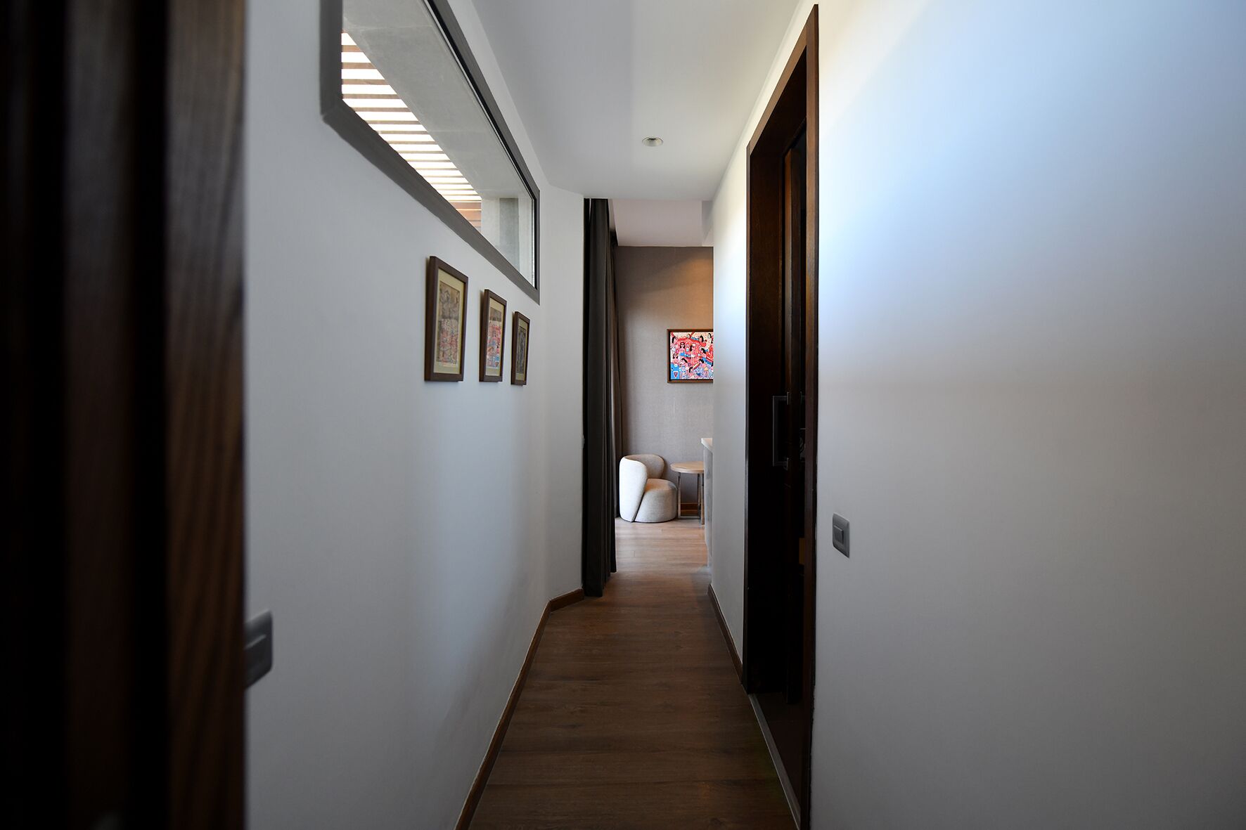 A hallway with wooden flooring, white walls, and framed artwork. There is a door on the right and a seating area in the distance.