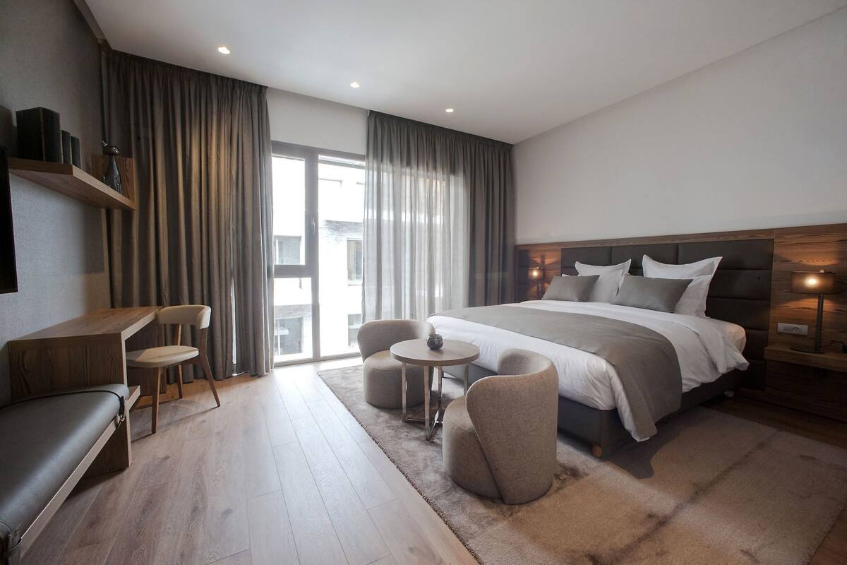 Une chambre d’hôtel moderne, dotée d’un grand lit, d’un bureau, de deux chaises, d’une petite table et offrant une vue sur l’extérieur.