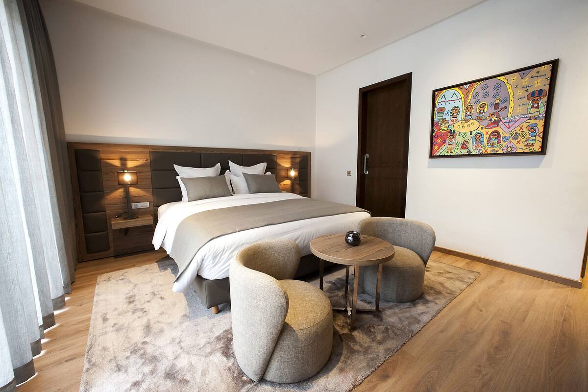 Une chambre d’hôtel moderne avec un grand lit, un fauteuil aux lignes arrondies, une petite table ronde et une œuvre d’art colorée accrochée au mur.