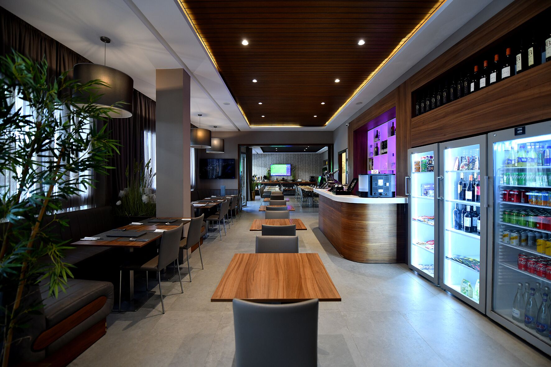A modern restaurant with wooden tables and chairs, a bar area, and a refrigerator stocked with beverages.