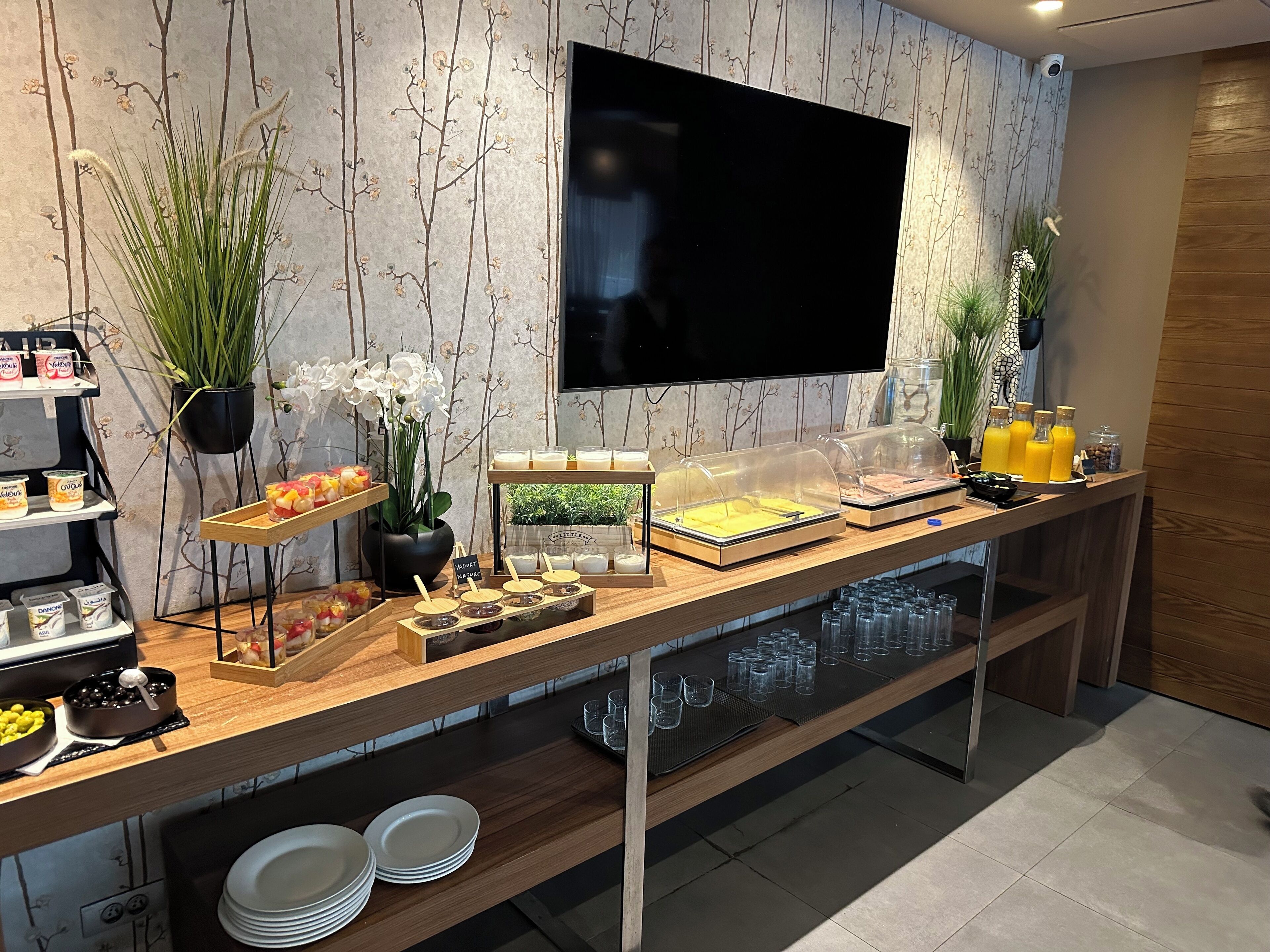 Un bufé de desayuno con diversas opciones de comida, un televisor montado en la pared y plantas decorativas.