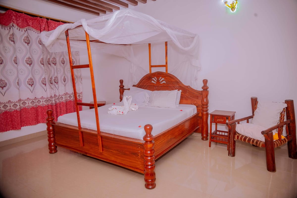 Una cama con dosel de madera, ropa de cama blanca y un dosel, una silla de madera y una mesita de noche junto a la cama.