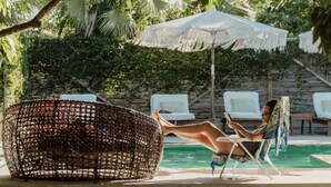 Un área junto a la piscina con una silla de mimbre, una tumbona y una sombrilla blanca.