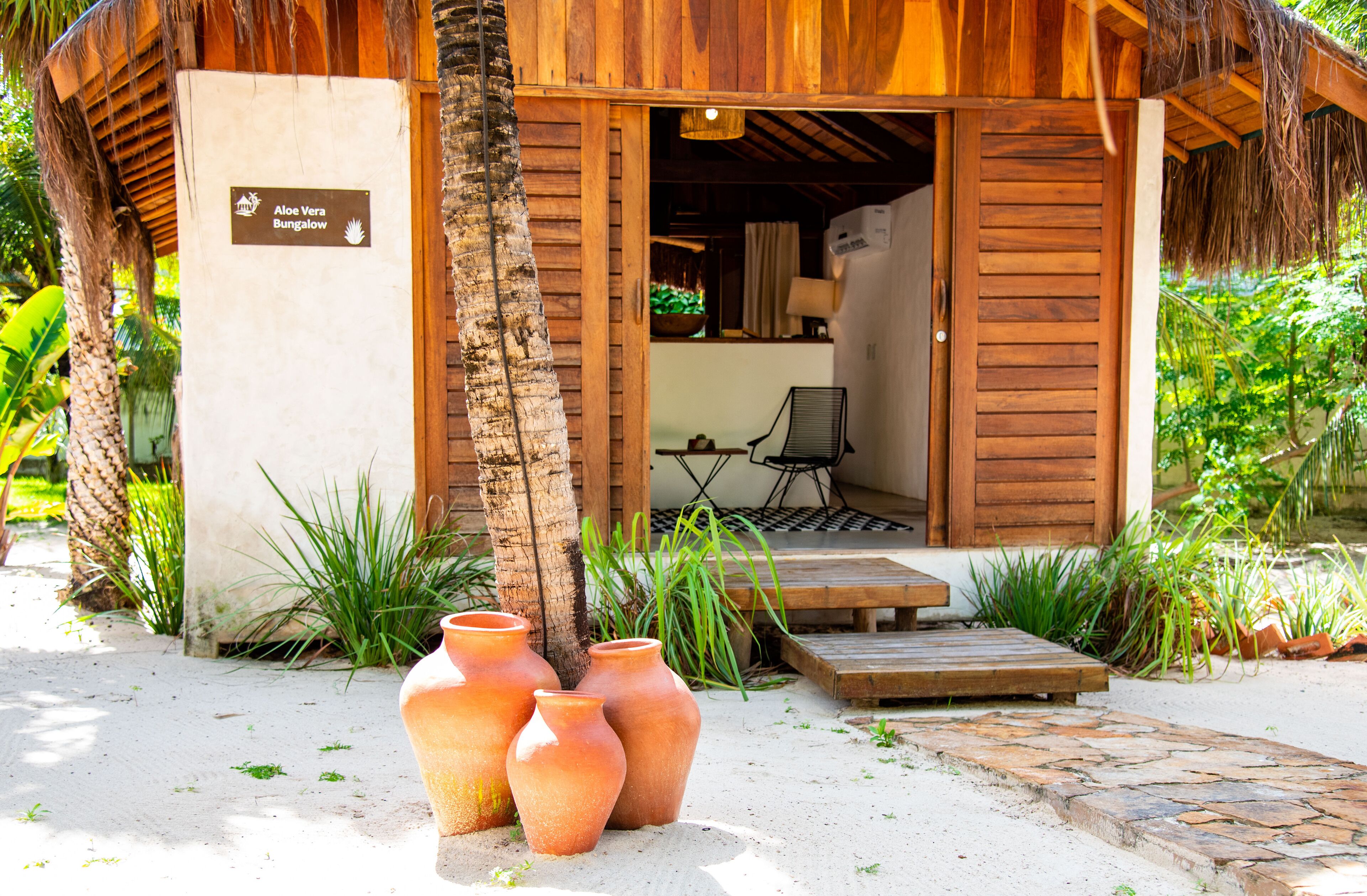 Una cabaña de madera con un letrero que dice "Aloe Vera Bungalow", un pequeño espacio de estar al aire libre y tres macetas grandes de terracota.