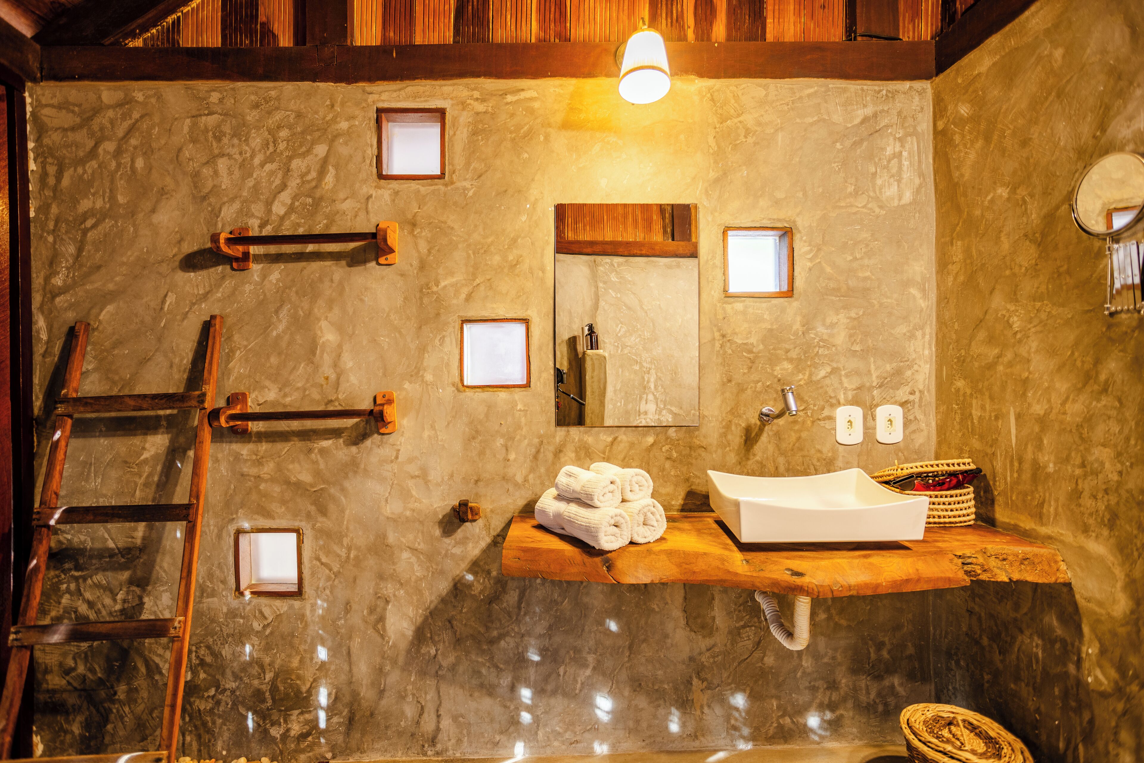 A bathroom with a stone wall, wooden ladder, a wooden shelf with towels, a sink, and a mirror.